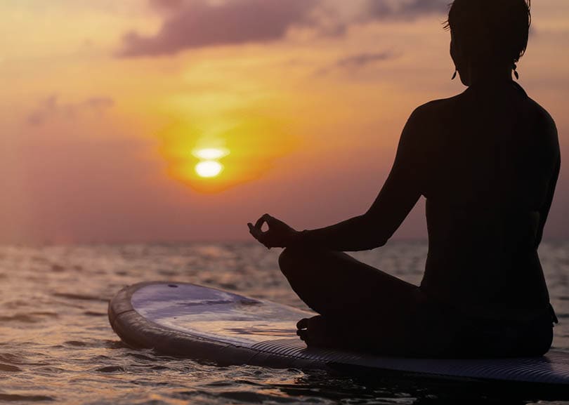 Woman practicing SUP yoga at sunset, meditating on a paddle board.