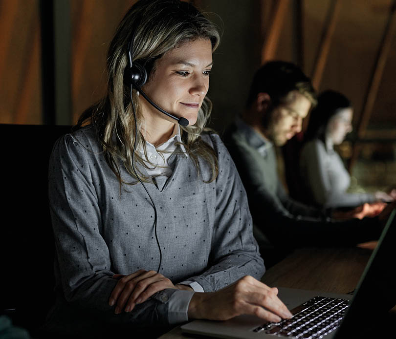 Group of entrepreneurs working on computers late in the office. Focus is on woman talking through hands-free device.